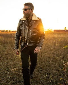 Full-length shot of a man wearing the shiny black Dawson rider jacket, styled with the removable cream shearling collar. The jacket is zipped up and worn outdoors/indoors.