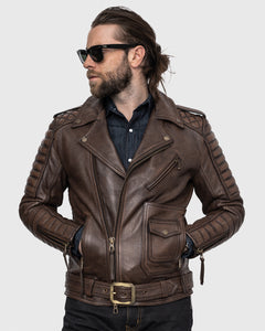 A person wearing a brown leather jacket stands against a white background. The jacket features antique-colored zippers and buckles, and a quilted pattern on the shoulders and elbows. The person is also wearing black sunglasses and is reflected in the shiny surface of the zipper. The image is taken from a side angle, showing the sleeves of the jacket hanging down